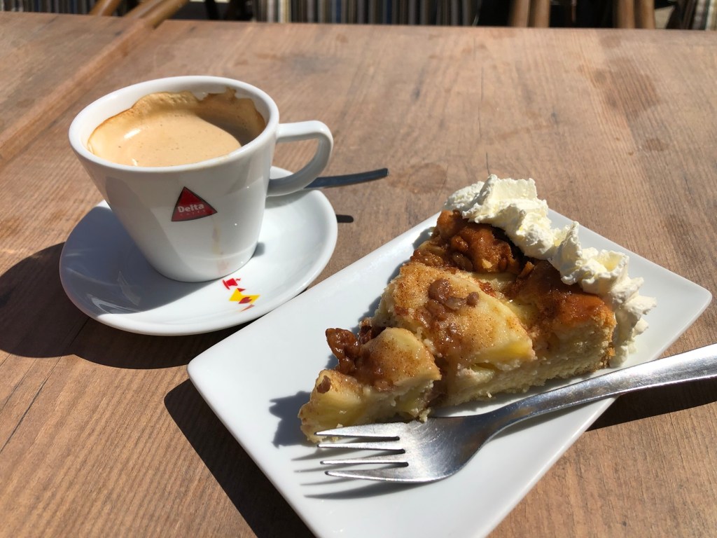 A cup of espresso with a saucer next to a slice of dessert topped with whipped cream on a white plate, resting on a wooden table.